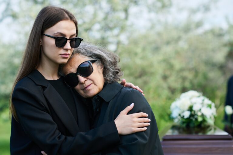Young woman in sunglasses and mourning attire supporting her grieving mother