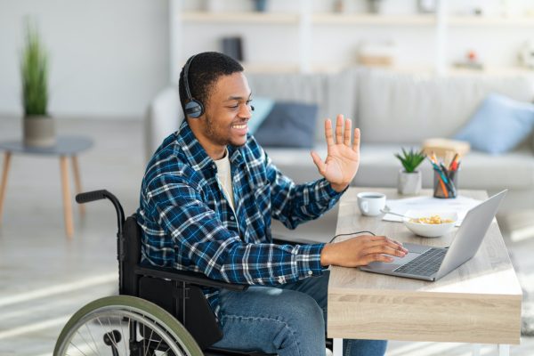 Happy young man in wheelchair communicating online, using laptop computer, waving at webcam at home